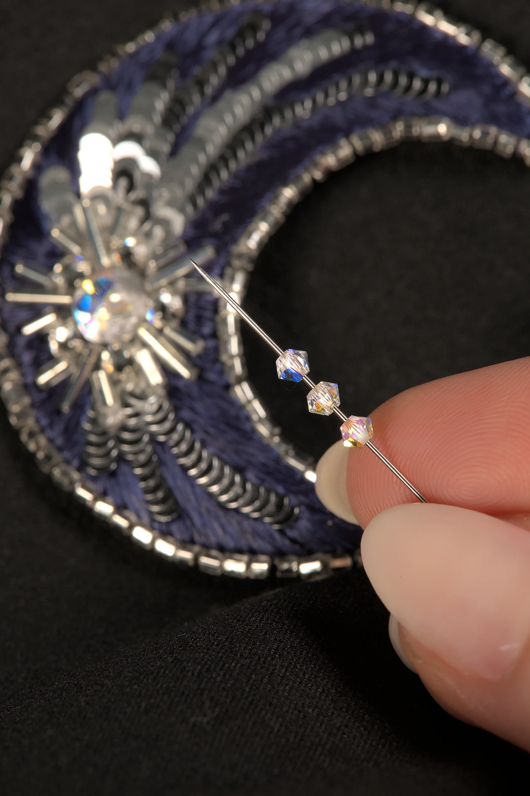 Primer plano de unos dedos sosteniendo una aguja con cristales de Swarovski, con un broche de luna bordado de fondo, mostrando un curso de bordado de Alta Costura en línea.