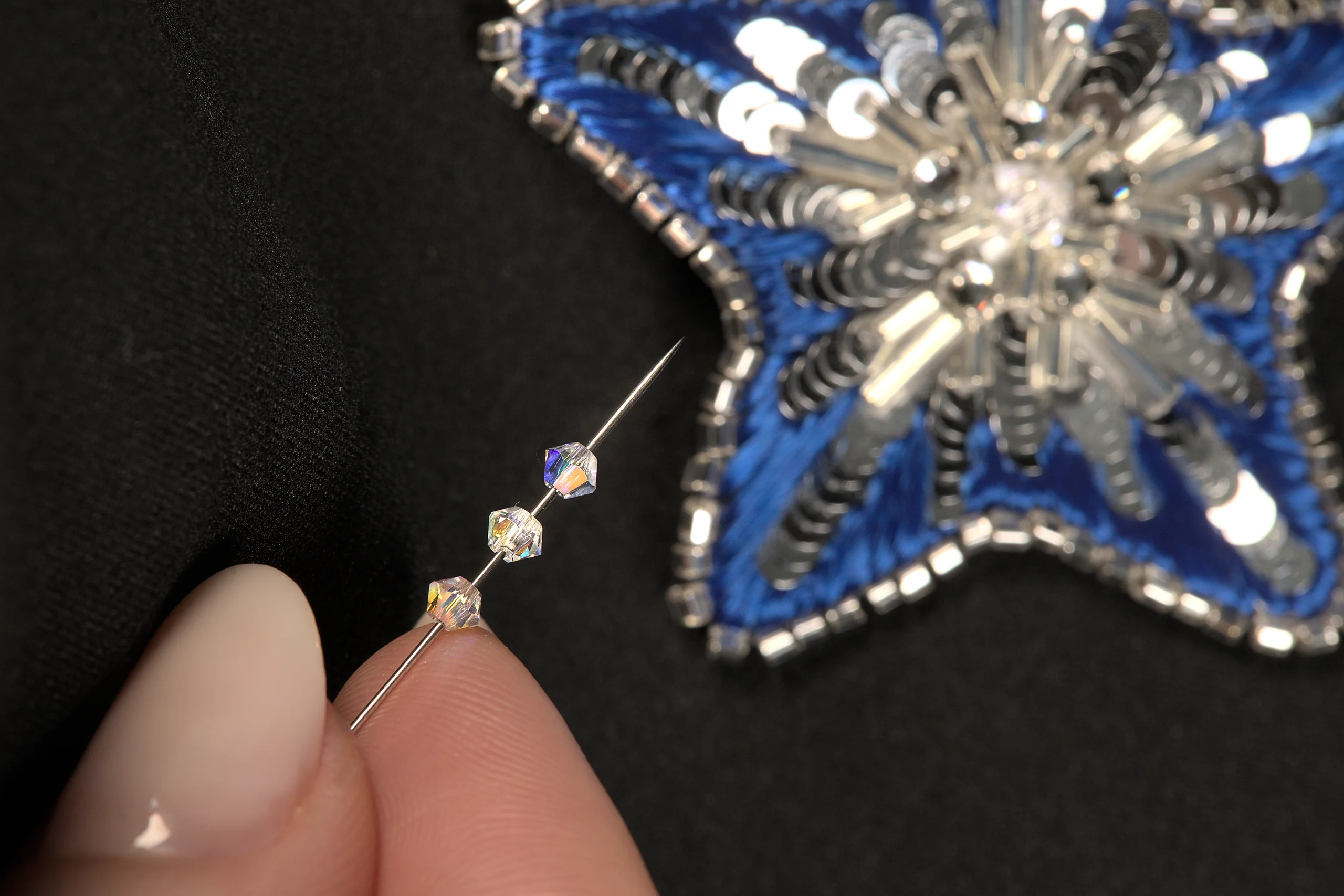 Close-up of fingers holding a needle with Swarovski crystals, with an embroidered star brooch in the background, showcasing an online Haute Couture embroidery course.