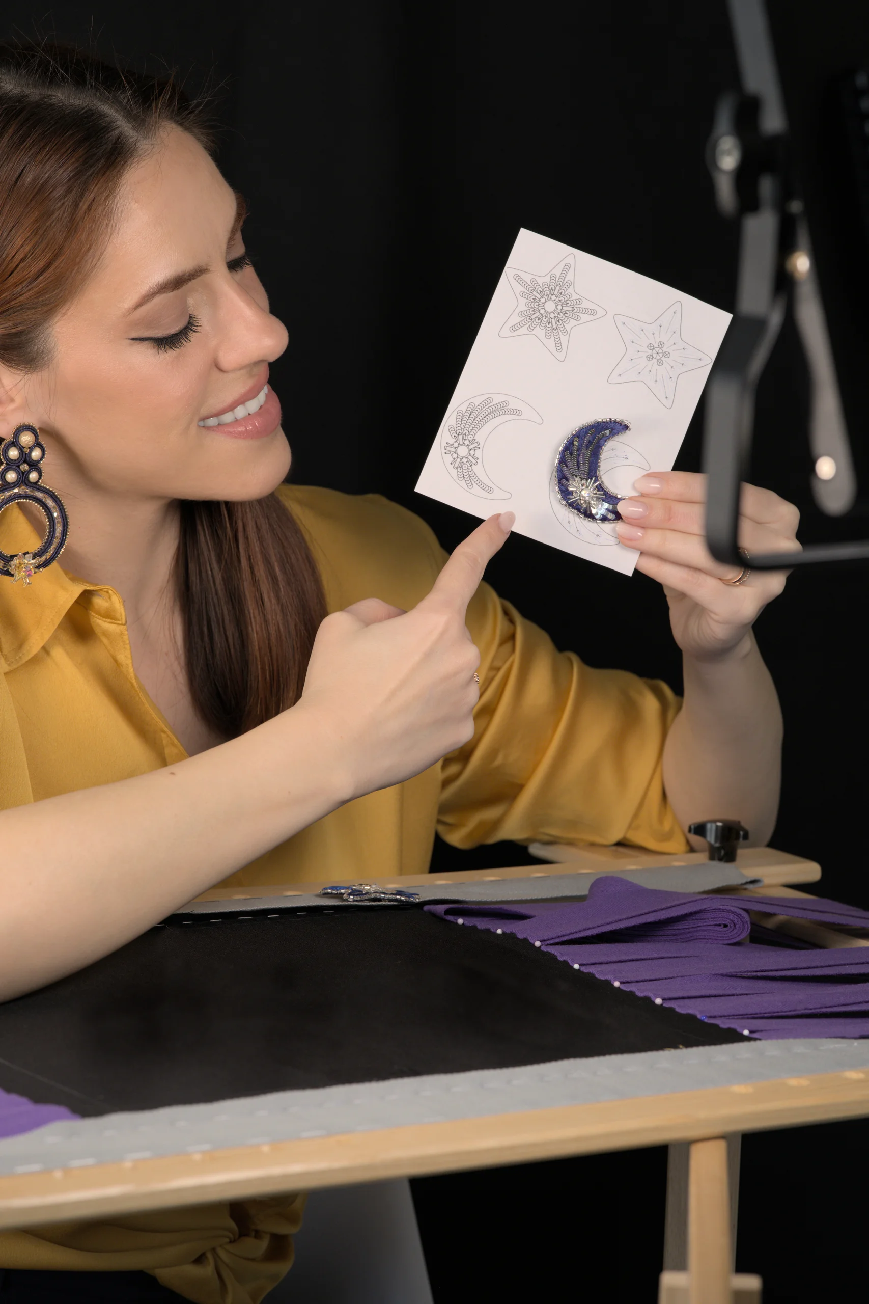 Laura S. Urbina showcases an embroidered moon brooch behind an embroidery frame, highlighting her online Haute Couture embroidery courses.